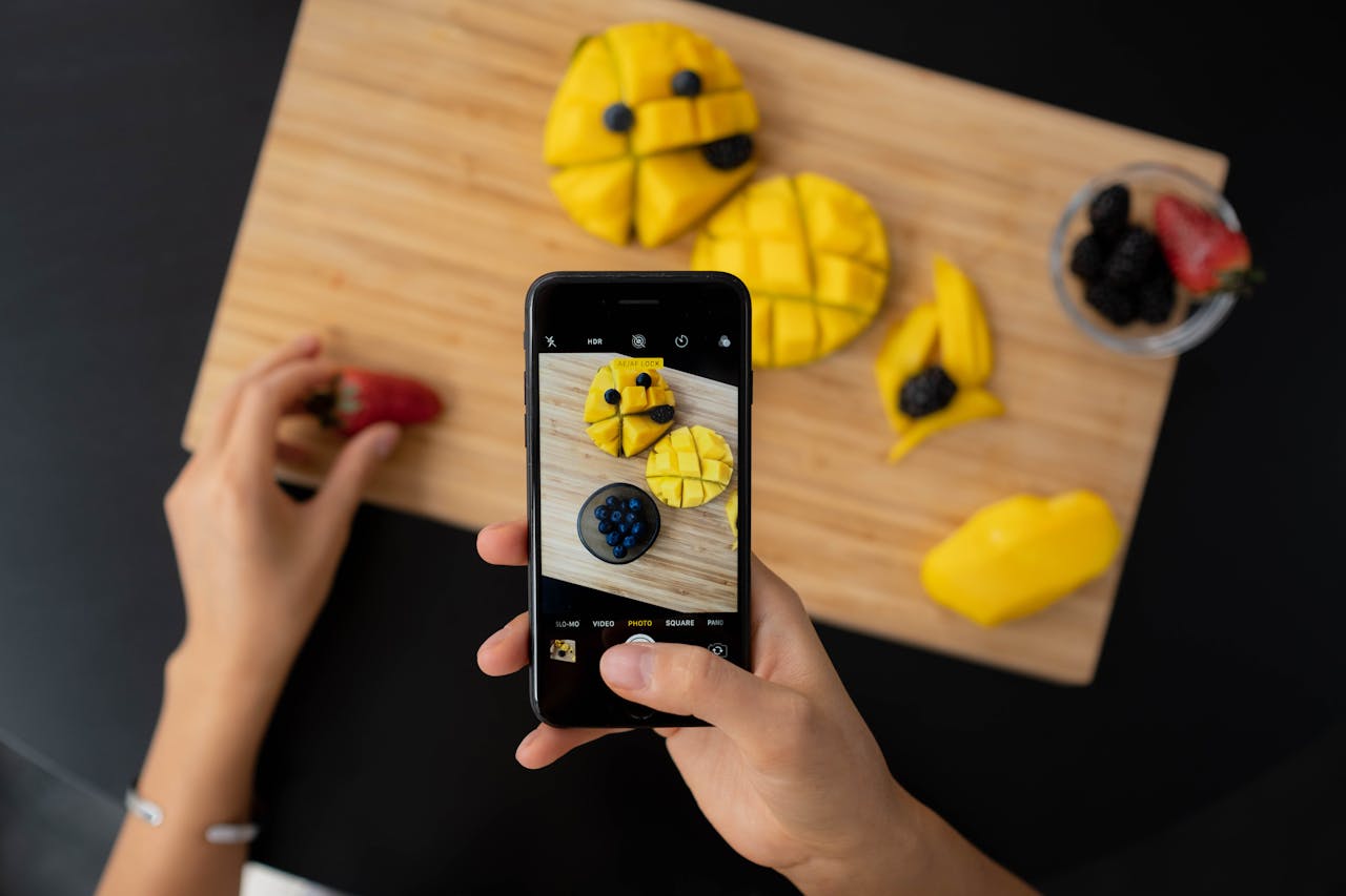 Overhead view of hands photographing fresh mango, strawberries, and blueberries with a smartphone on a cutting board.